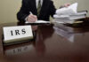 IRS sign on desk, person handling paperwork behind