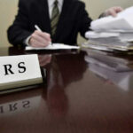 IRS Chaos: 34,000 Workers Cut IRS sign on desk, person handling paperwork behind