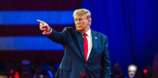 Man in suit pointing on stage with audience behind.