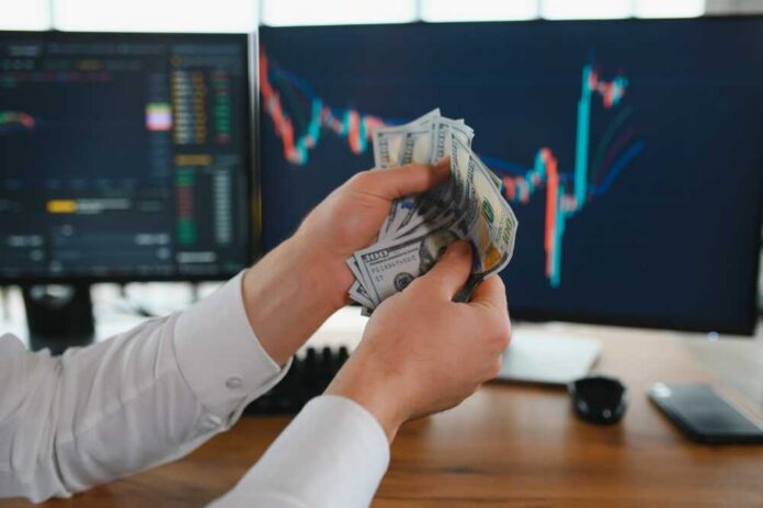 Person holding cash in front of stock market charts on computer screens