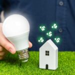 A person holding a light bulb next to a small house model with dollar signs