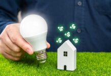 A person holding a light bulb next to a small house model with dollar signs