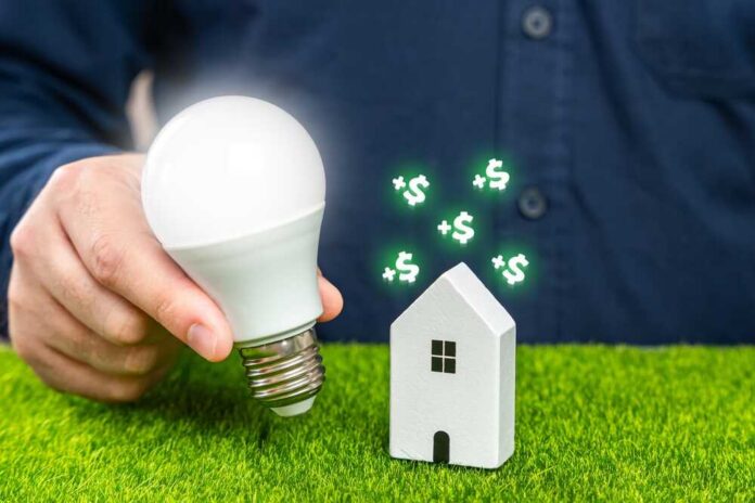A person holding a light bulb next to a small house model with dollar signs