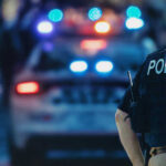 Teen Takeover ALERT: Curfew Ordered Police officer stands near patrol car with lights flashing.