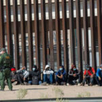 Arrest Quota? ICE Numbers Explode Border patrol officer watches group sitting by a fence.