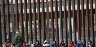 Border patrol officer watches group sitting by a fence.