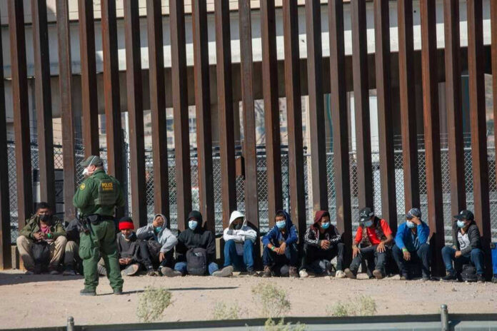 Border patrol officer watches group sitting by a fence.