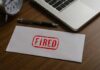Fired By Trump, Then Promoted?! An envelope marked 'FIRED' on a wooden desk next to a clock and a laptop