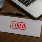Fired By Trump, Then Promoted?! An envelope marked 'FIRED' on a wooden desk next to a clock and a laptop