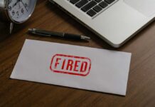 Fired By Trump, Then Promoted?! An envelope marked 'FIRED' on a wooden desk next to a clock and a laptop