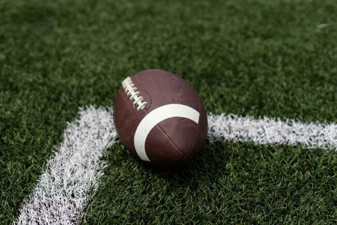 Close,Up,View,Of,An,American,Football,Sitting,On,A A brown football resting on green artificial turf near a white line