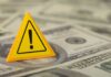 A yellow warning sign placed on a pile of dollar bills