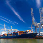 NEW: Hormuz Explodes, Tankers Hit, Blockade Back Cargo ship docked at port with cranes unloading.