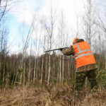 Hunter’s Shot Kills Mom – ‘Thought It Was Deer’ Hunter in high-visibility vest aiming rifle in forest.