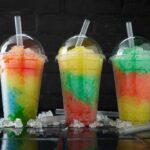 Three colorful shaved ice drinks in clear plastic cups with domed lids