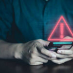 Beloved Mascot Stops Laughing – Why Now? Person holding phone with warning symbol displayed.