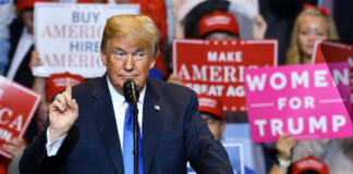Donald Trump speaks at a rally with Bill Kenney in the crowd holding signs