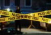 MURDER: Detective Charged Police tape marking a crime scene with blurred figures in the background