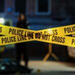 MURDER: Detective Charged Police tape marking a crime scene with blurred figures in the background