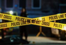 MURDER: Detective Charged Police tape marking a crime scene with blurred figures in the background