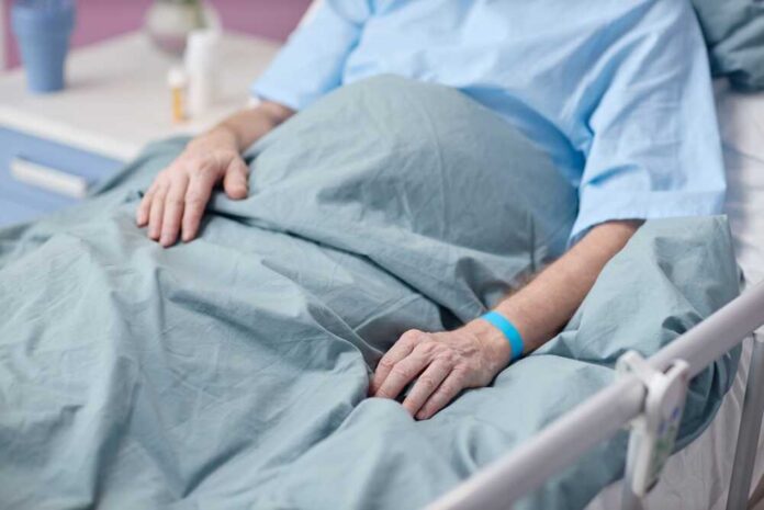 Senior,Caucasian,Man,Lying,In,Hospital,Bed,With,Medical,Wristband Patient resting in a hospital bed with hands on blanket
